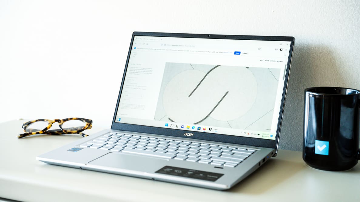 An open and powered on silver laptop sitting on a white surface with a black mug on the right and a pair of glasses on the left