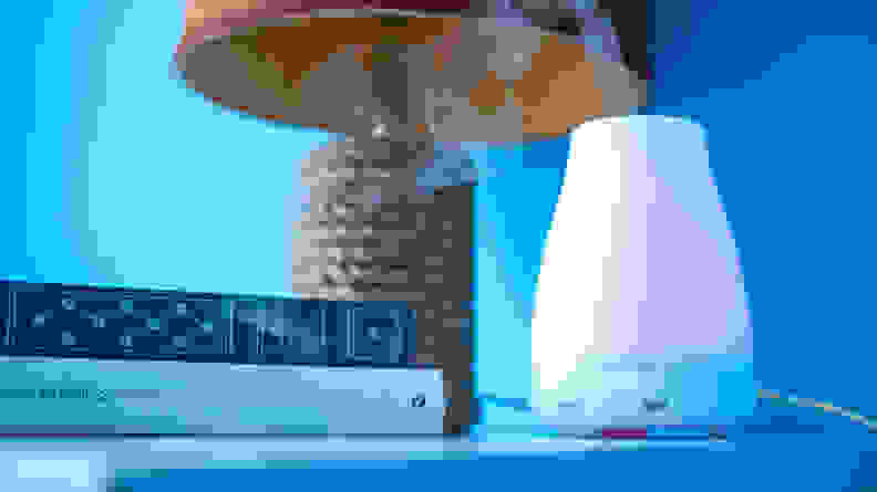 A white, conical InnoGear essential oil diffuser emitted vapor on a table with books and a lamp. The picture has a blue cast.