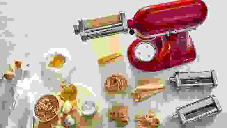 KitchenAid pasta attachment rolling pasta dough on a marble surface, surrounded by pasta ingredients