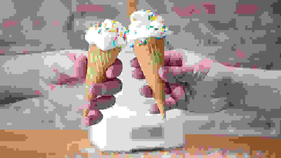 Two hands holding ice cream cones with vanilla ice cream and rainbow sprinkles cheers in front of an ice cream maker on a wood counter in front of an exposed brick wall.
