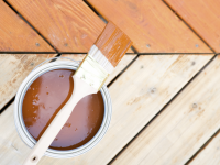 Deck boards, half of which have been stained. On the decking sits an open can of stain and a paintbrush.