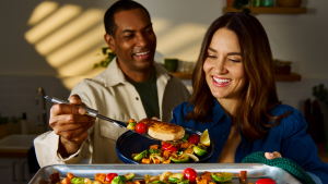 A couple smiles while plating a HelloFresh meal in a bright kitchen, showcasing the joy of cooking with a HelloFresh promo code.