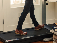 Close-up of a black under-desk treadmill in use at a standing desk in the corner of the room. There is a stack of hardcover books beside it.